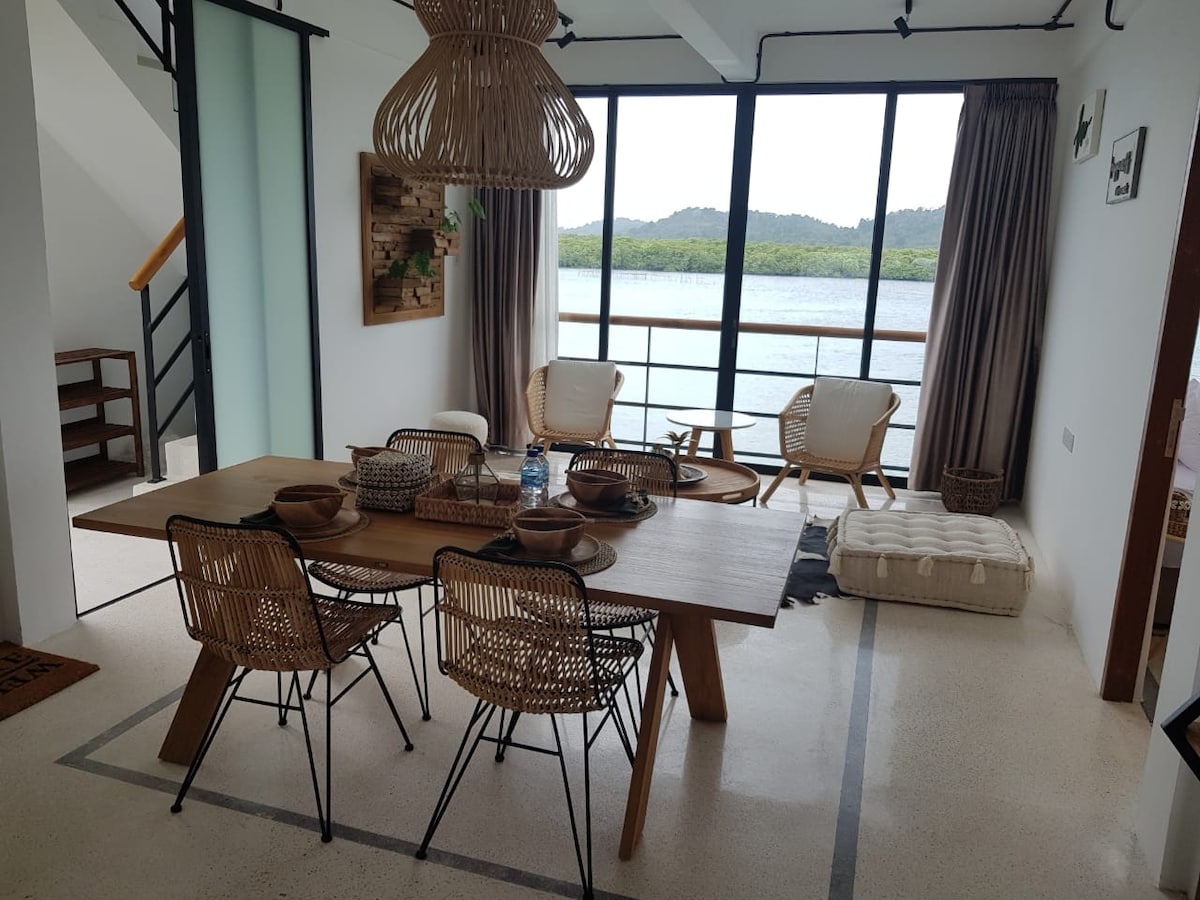 A dining area features a wooden table surrounded by six rattan chairs, complemented by a cushion seating area. Large windows provide a view of the landscape, while light drapes softly frame the windows. A staircase is visible, enhancing the open layout.