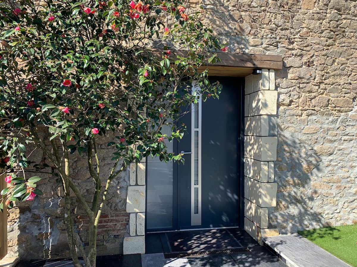 A modern entrance door is framed by a stone wall, featuring a stylish design with vertical panels. A flowering shrub adds a touch of greenery beside the door, enhancing the welcoming ambiance of the entryway.