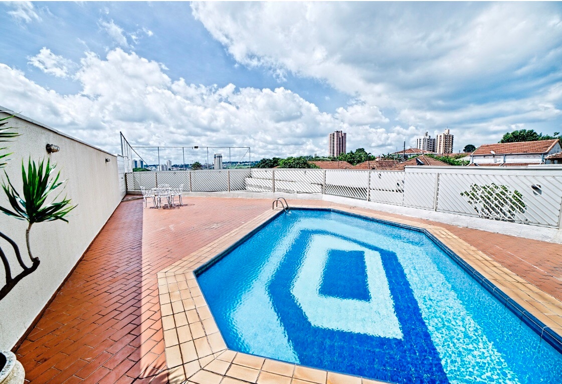 A rooftop pool area features a geometric-shaped pool accentuated by tiled patterns. Surrounding spaces include a tiled deck with lounge chairs, offering scenic views of the skyline and partially cloudy skies. Fencing provides privacy while creating an inviting setting for relaxation.