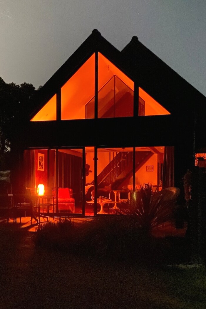 A modern wooden cottage is illuminated in warm orange light against the night sky. Large glass windows reveal a cozy interior, featuring a staircase and seating area, while outdoor seating creates an inviting atmosphere.