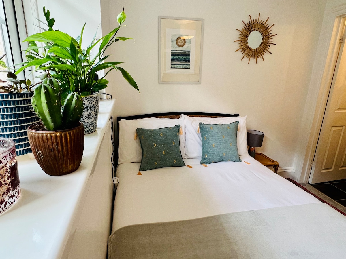 A cozy bedroom features a spacious bed adorned with two decorative pillows. A plant sits on the window ledge, complemented by a wall-mounted art piece. Natural light gently illuminates the room, highlighting the neutral tones and tasteful décor.