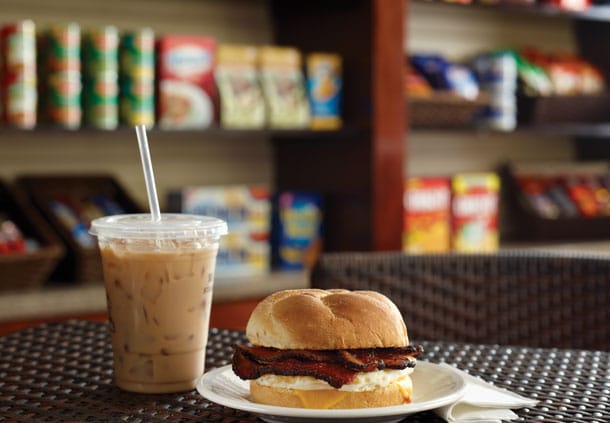 A freshly made sandwich is presented on a white plate alongside a clear cup of iced coffee with a straw. In the background, shelves filled with snacks and beverages are organized for easy access.
