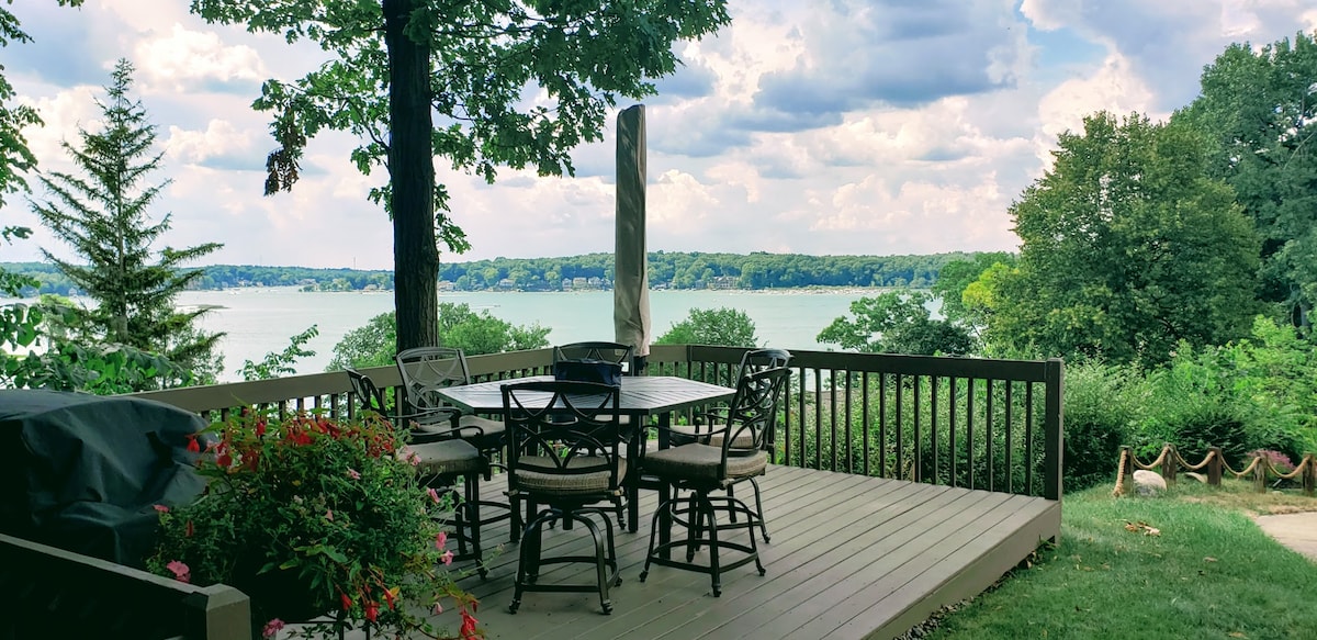 An outdoor deck features a large dining table surrounded by six chairs, situated under the shade of a tree. Lush green landscape and a serene lake view are visible in the background, offering a calm and spacious environment for outdoor dining.