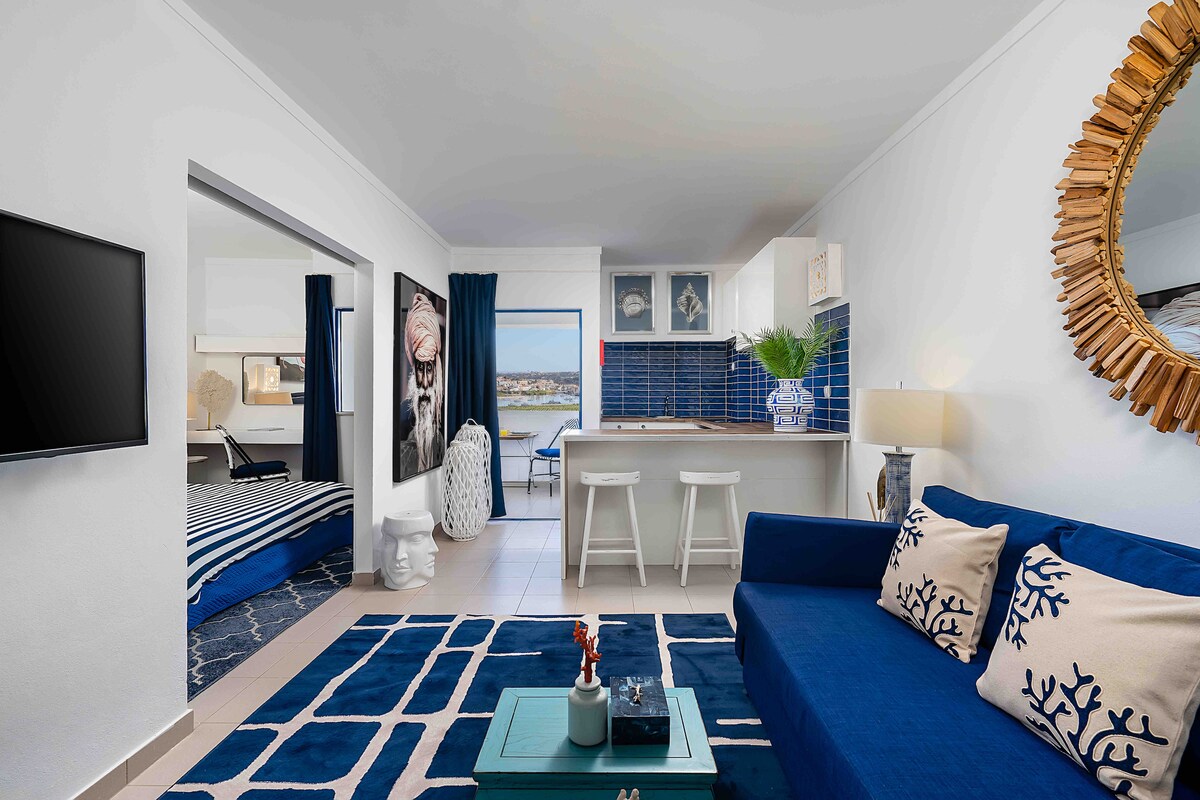 A bright living area is featured, with a blue sofa complemented by decorative cushions. The open kitchen with light cabinetry is visible in the background, along with two bar stools. Natural light enters through a large window, enhancing the coastal aesthetic of the space.