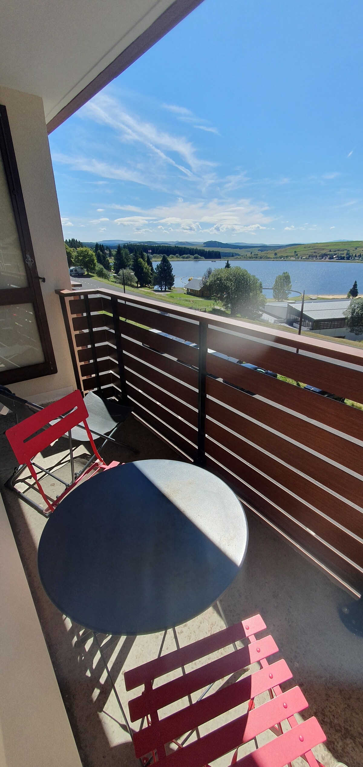 A balcony is featured, overlooking a serene body of water under a clear blue sky. Two red chairs and a small round table are positioned within the space, providing an inviting area for relaxation. The balcony's railing enhances the open view.