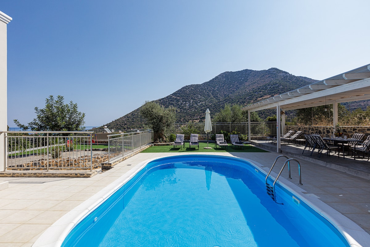 A private swimming pool is set in a serene outdoor area, surrounded by comfortable sunbeds. The scenic backdrop features lush greenery and a mountain. A shaded dining area with a large table is available, providing a perfect space for outdoor meals.