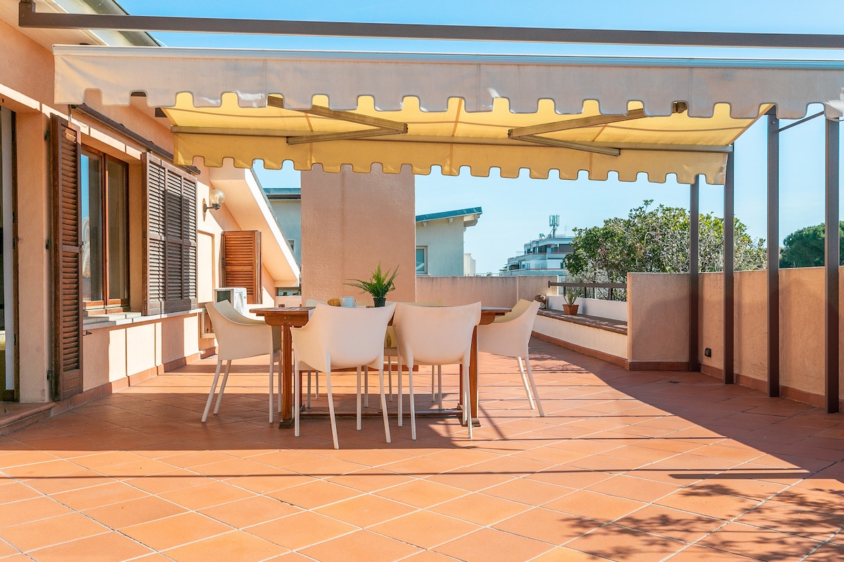 An outdoor terrace features a shaded dining area with a rectangular table surrounded by several white chairs. The space is defined by terracotta tiles and is framed by a sunshade that offers protection from the elements. Surrounding greenery adds a touch of nature.