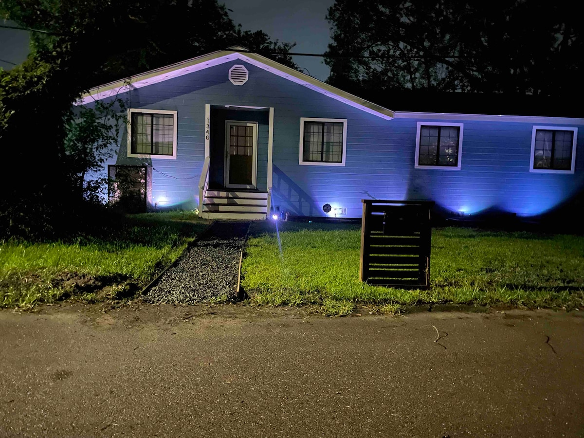 The exterior of a renovated blue home is illuminated by soft lighting at night. A clear path leads to the front door, framed by lush greenery. Nearby, a decorative mailbox is positioned on the grass, contributing to the inviting entrance.