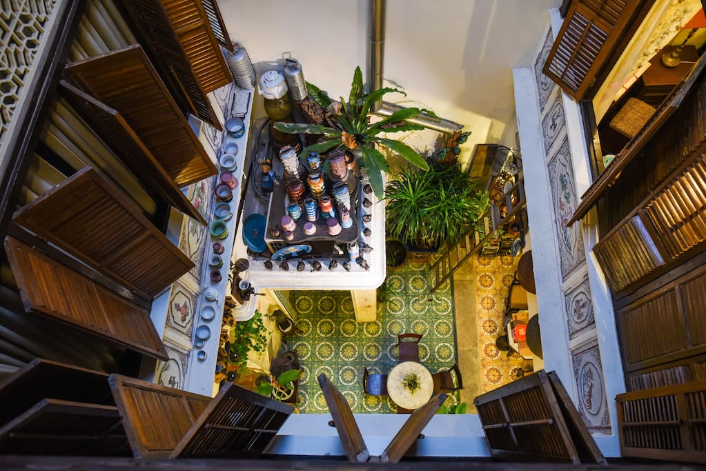 An overhead view reveals a vibrant common area adorned with greenery. A central countertop displays various colorful jars, surrounded by intricate tile flooring. Wooden shutters frame the scene, enhancing the airy ambiance of this inviting space.