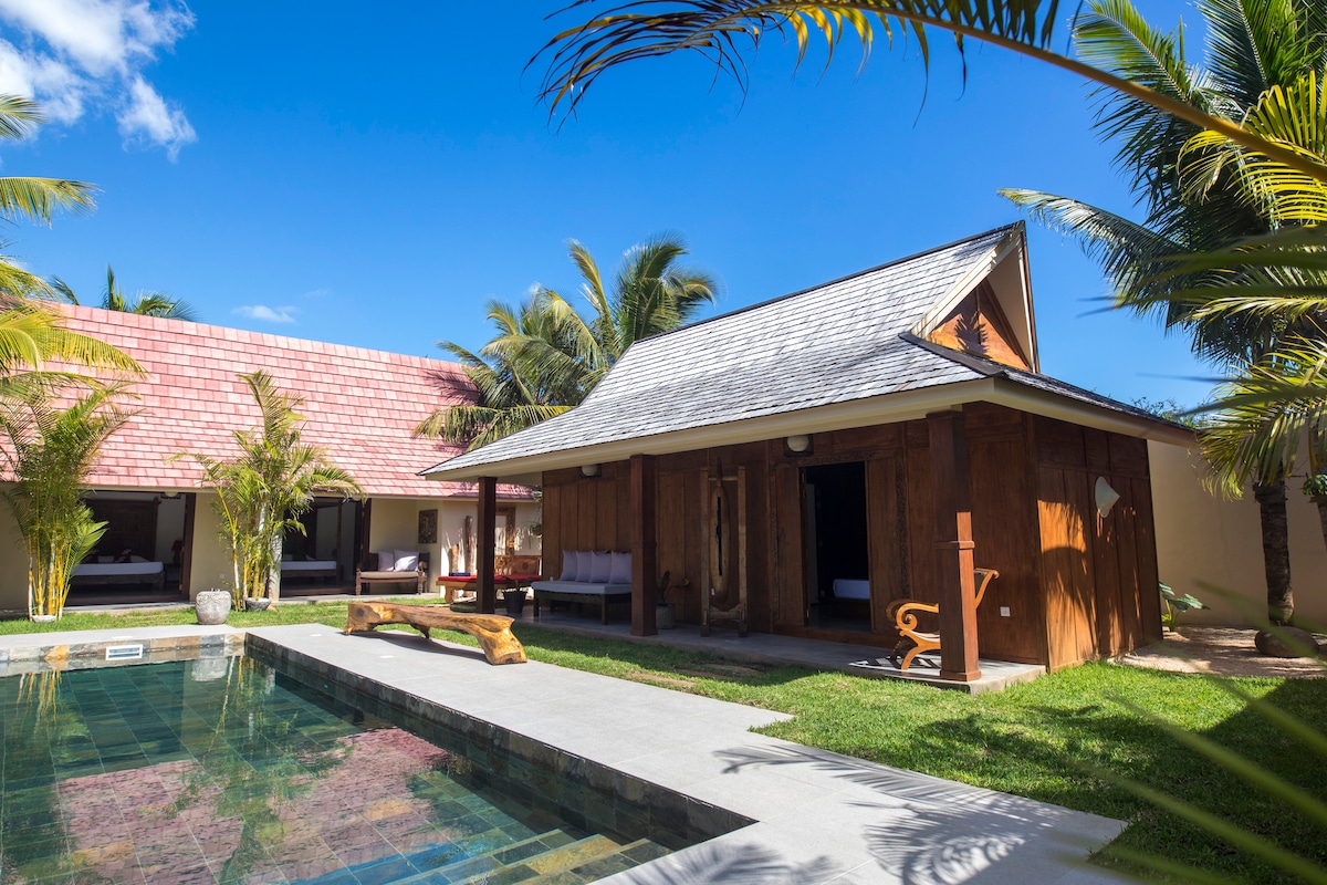 The villa showcases a traditional Javanese architectural style with wooden features. The inviting pool reflects sunlight, surrounded by lush tropical vegetation and palm trees. Comfortable outdoor seating is available, creating a serene outdoor environment for relaxation.
