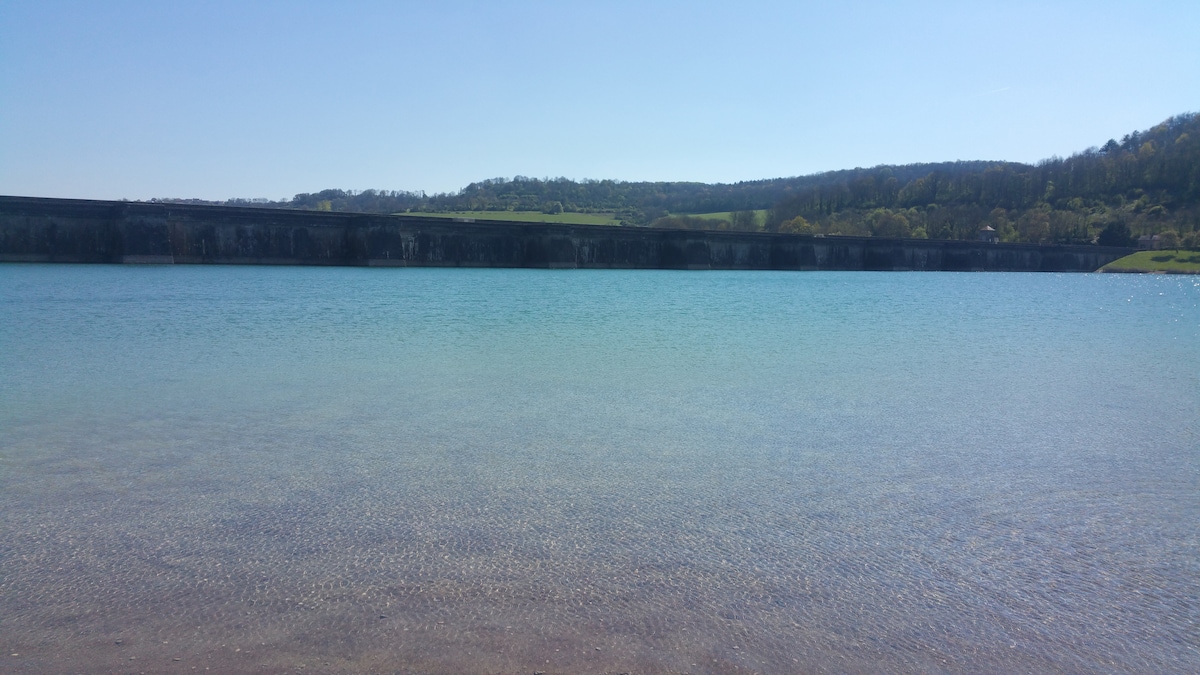 A calm body of water is showcased, with a smooth sandy shoreline visible in the foreground. The water exhibits a clear turquoise hue, reflecting the sunlight. In the background, a stone structure lines the water's edge, accompanied by gently rolling hills of greenery.