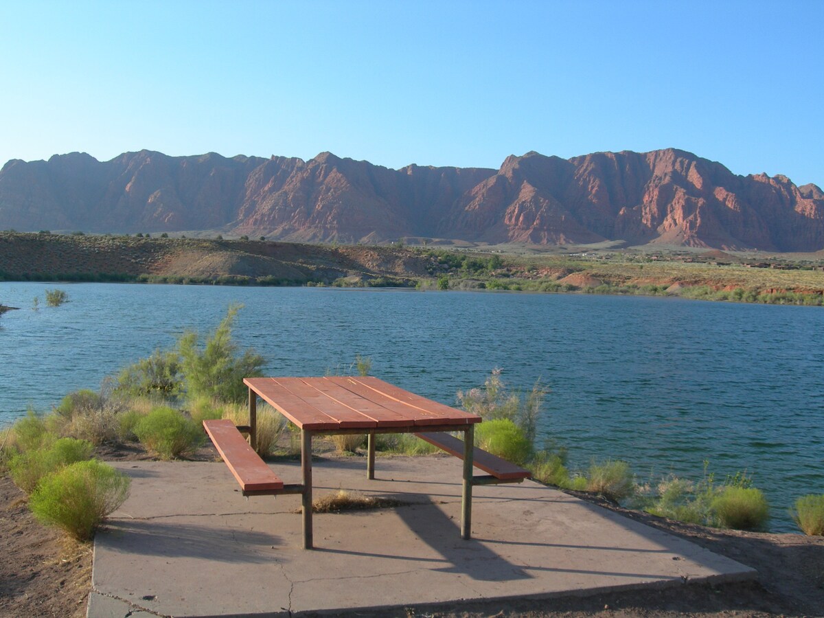 A picnic table made of wood sits on a concrete pad near a serene body of water. Lush green shrubs frame the scene, with striking red rock formations rising majestically in the background under a clear blue sky.