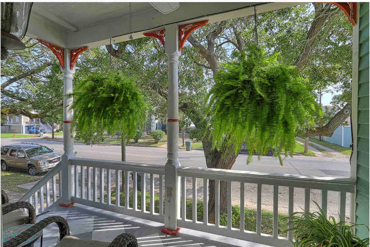 A front porch is adorned with hanging ferns, creating a lush ambiance. The space features a railing painted in white, with ornamental columns in red. A peaceful street view is visible, framed by leafy trees, enhancing the tranquil atmosphere.