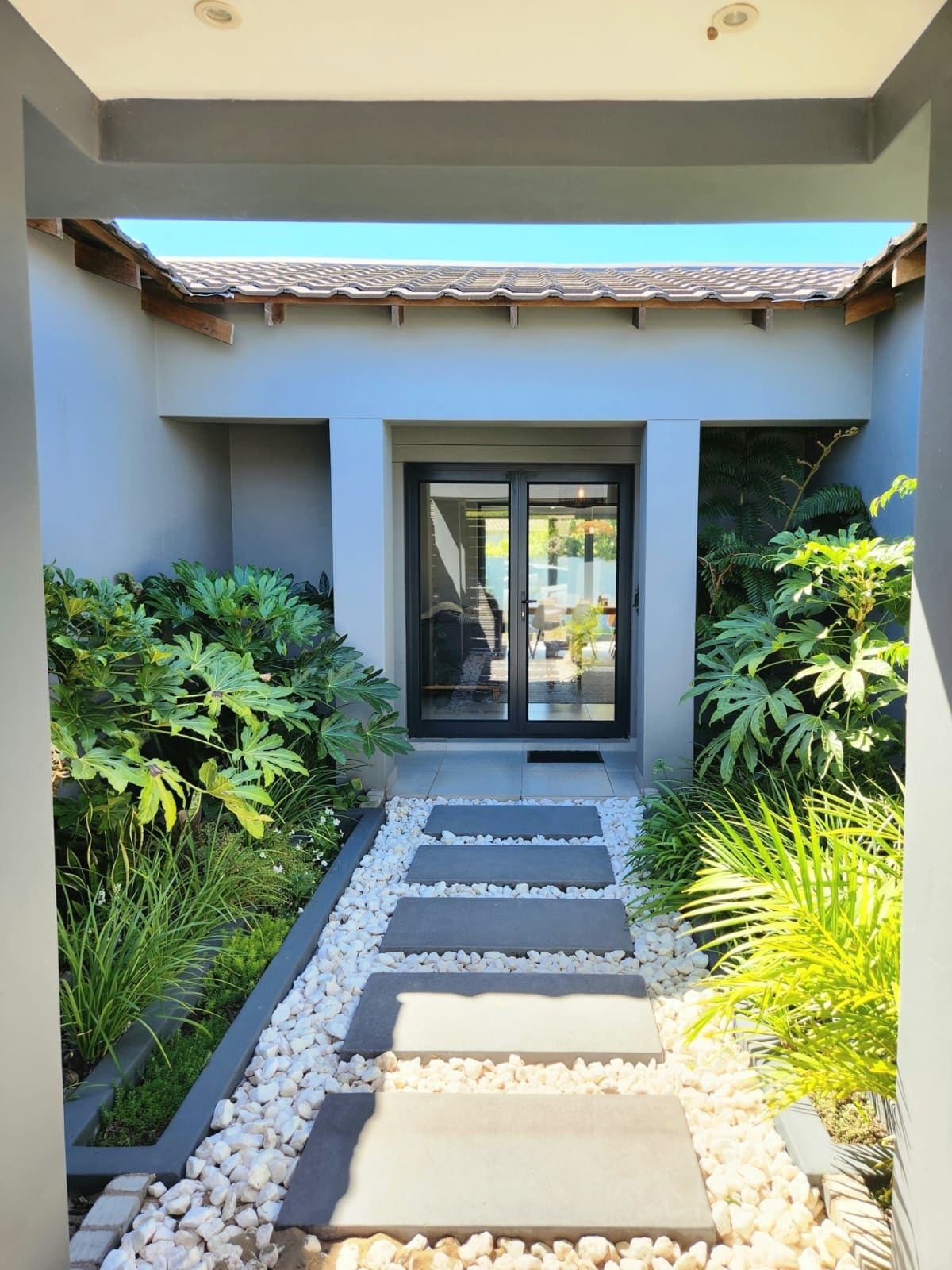 A stone pathway bordered by lush greenery leads toward a double glass door entrance. The surrounding plants provide a vibrant touch, enhancing the tranquil atmosphere of the entrance. Sunlight streams through, illuminating the inviting space ahead.