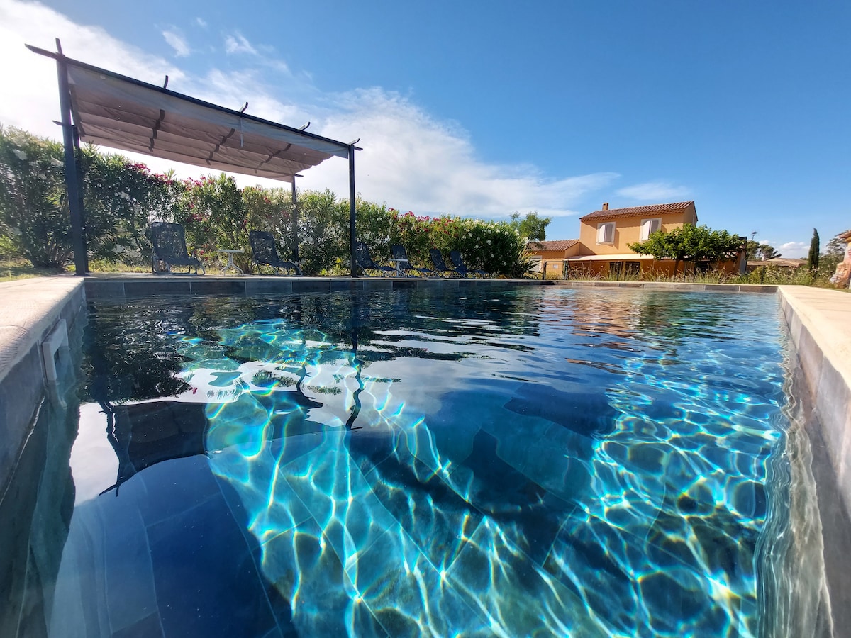 The private swimming pool reflects the clear blue sky and features a spacious deck area with sun loungers. Lush greenery surrounds the pool, creating a serene setting. A shaded gazebo is visible in the background, providing an inviting spot for relaxation.