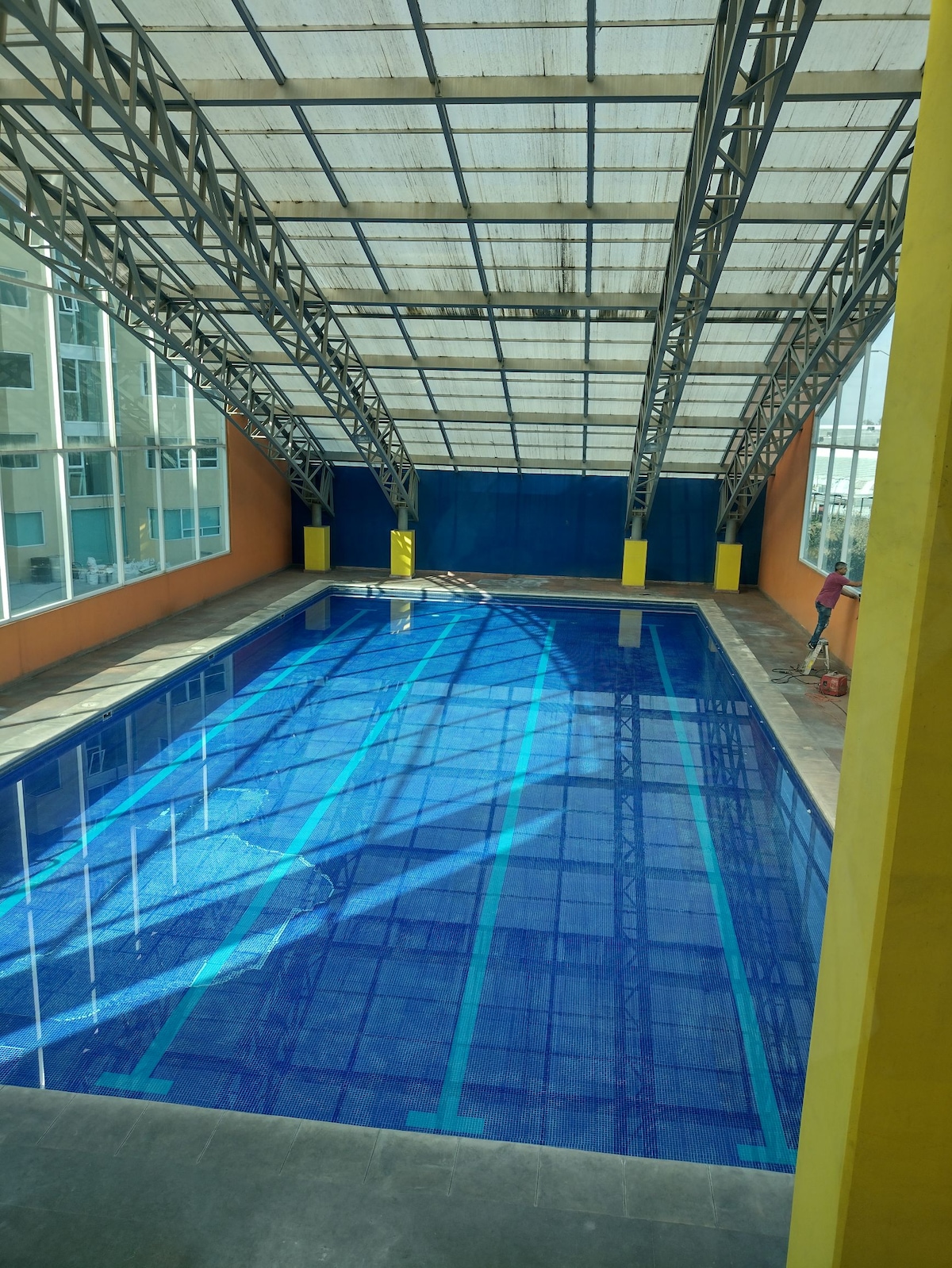 An indoor swimming pool is visible, featuring a vibrant blue surface and clear water. The pool is enclosed within a structured frame, with transparent roofing allowing natural light to illuminate the area. Yellow support columns accentuate the space, contributing to a bright environment.