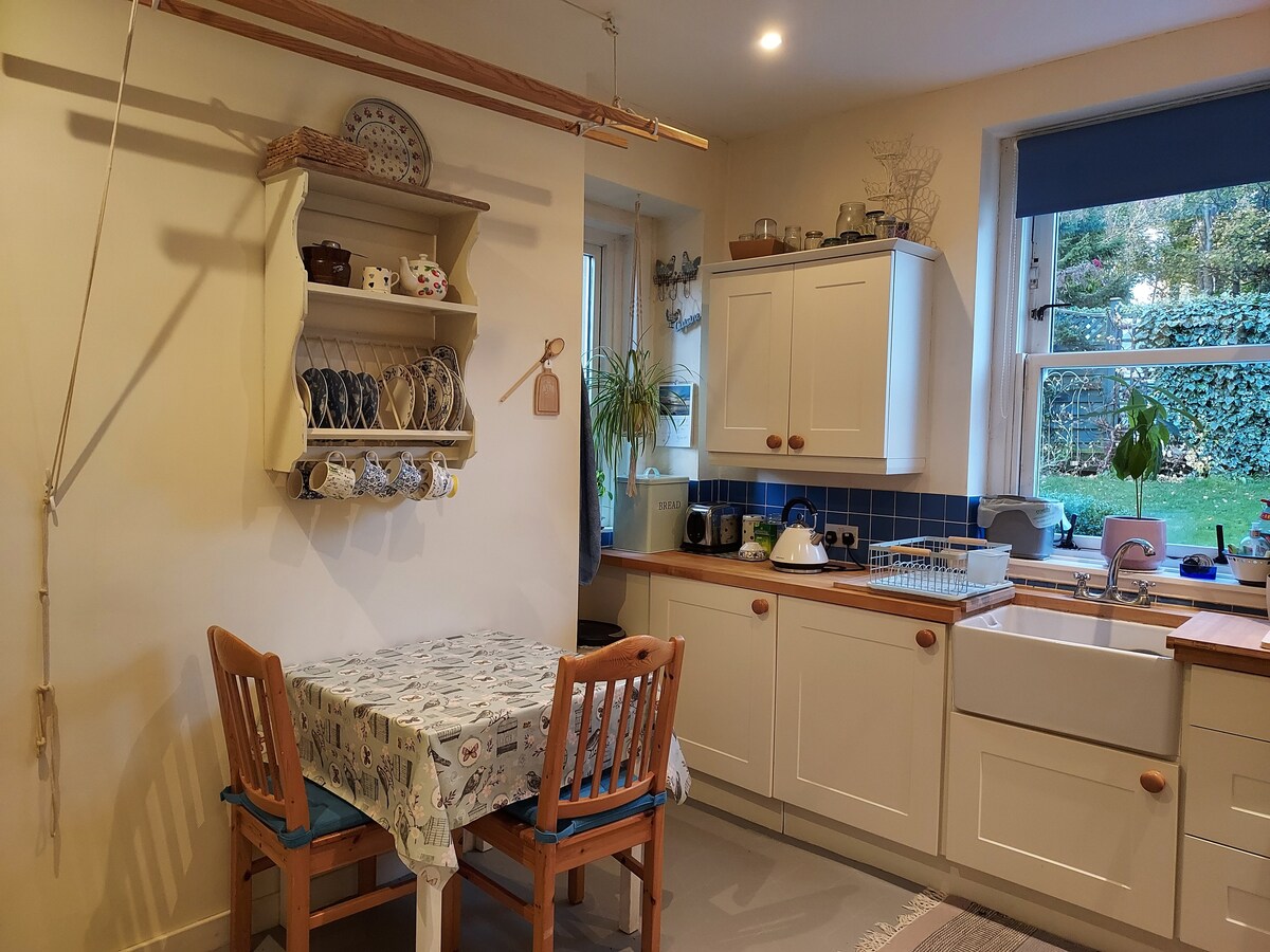 A functional kitchen area includes a small table with two wooden chairs, topped with a patterned cloth. Shelves display dishware and cups. Natural light enters through a window overlooking the garden, while a bowl and kettle sit on a countertop beside a sink.