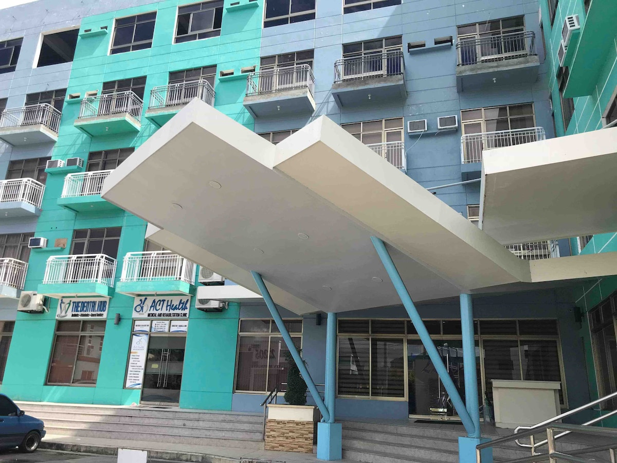 The exterior of the condominium building is captured, showcasing a modern design with a prominent overhang at the entrance. Brightly colored walls in shades of turquoise are complemented by balconies on the upper levels, providing a cheerful and welcoming appearance.