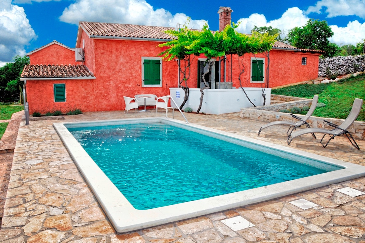 A self-standing house features a vibrant coral exterior complemented by green window shutters. A private swimming pool, surrounded by a stone patio, is positioned in front of the house. Sun loungers are placed nearby, with a lush green garden visible in the background.