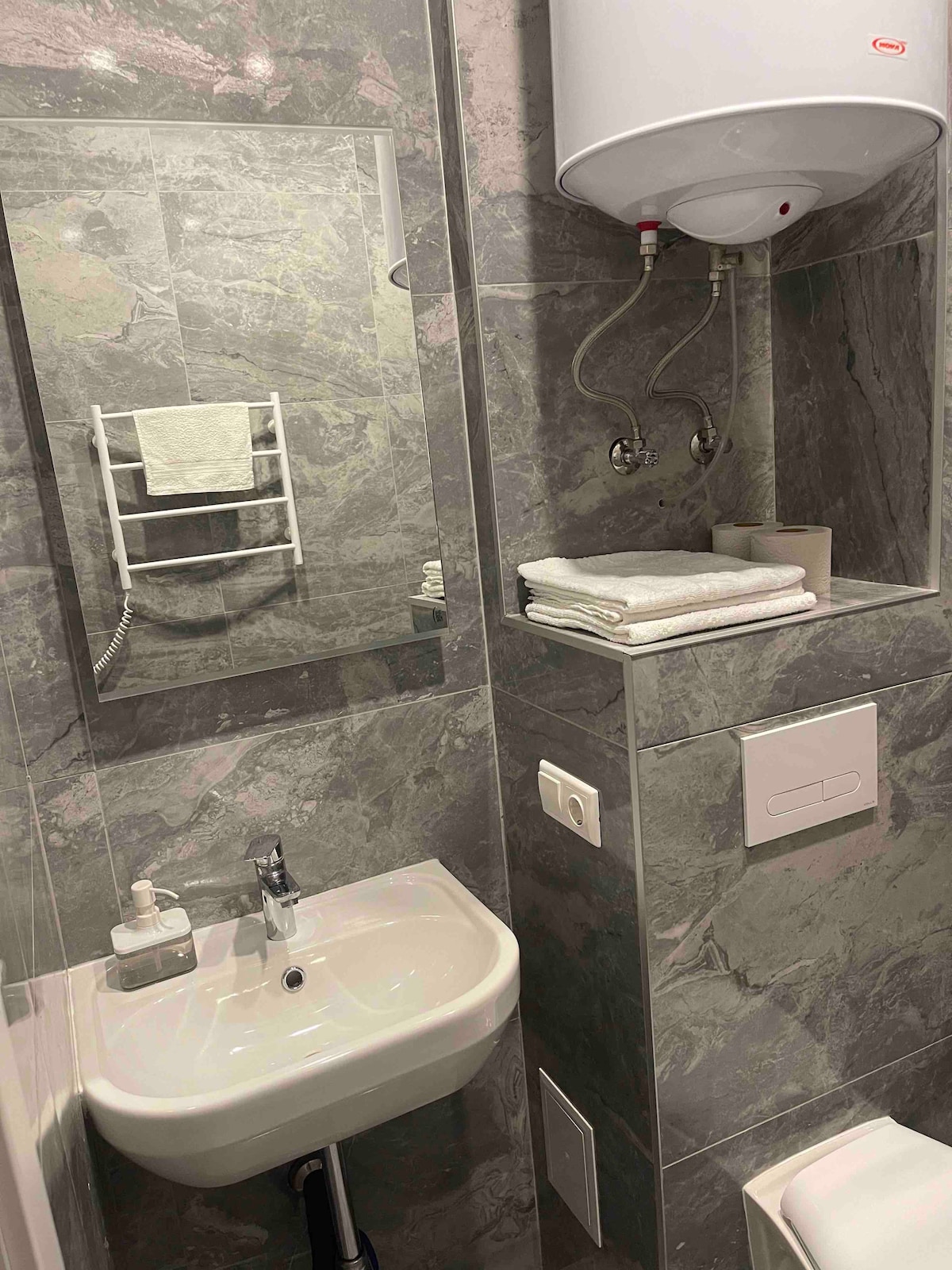 A compact bathroom features a sleek sink set against stylish gray marble walls. Soft, folded towels are neatly arranged on a shelf. A mirror with a simple frame reflects the well-maintained fixtures, while a hairdryer is conveniently hung nearby.