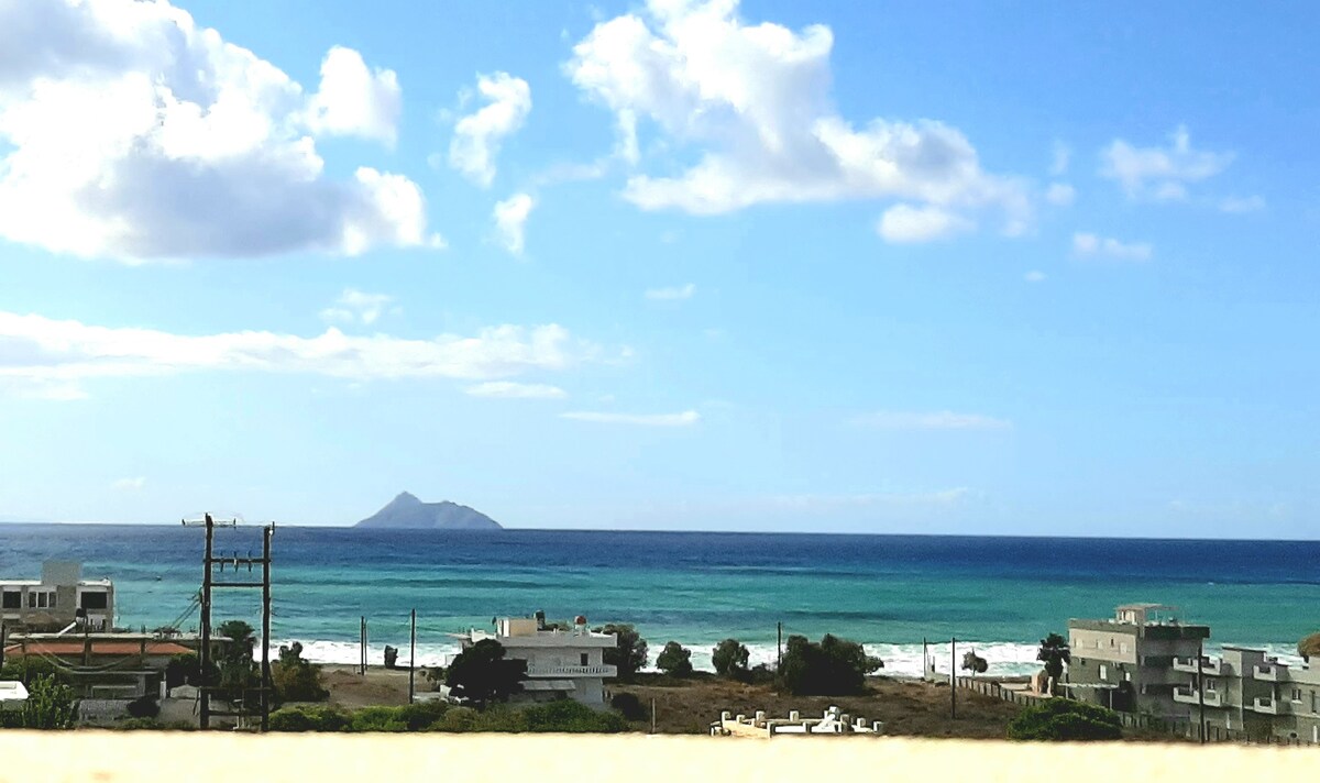 A panoramic view of the sea is captured, showcasing varying shades of blue and green waves. The horizon features distant islands, while the foreground includes coastal villages with houses. Fluffy white clouds drift across the sky, enhancing this tranquil seaside landscape.