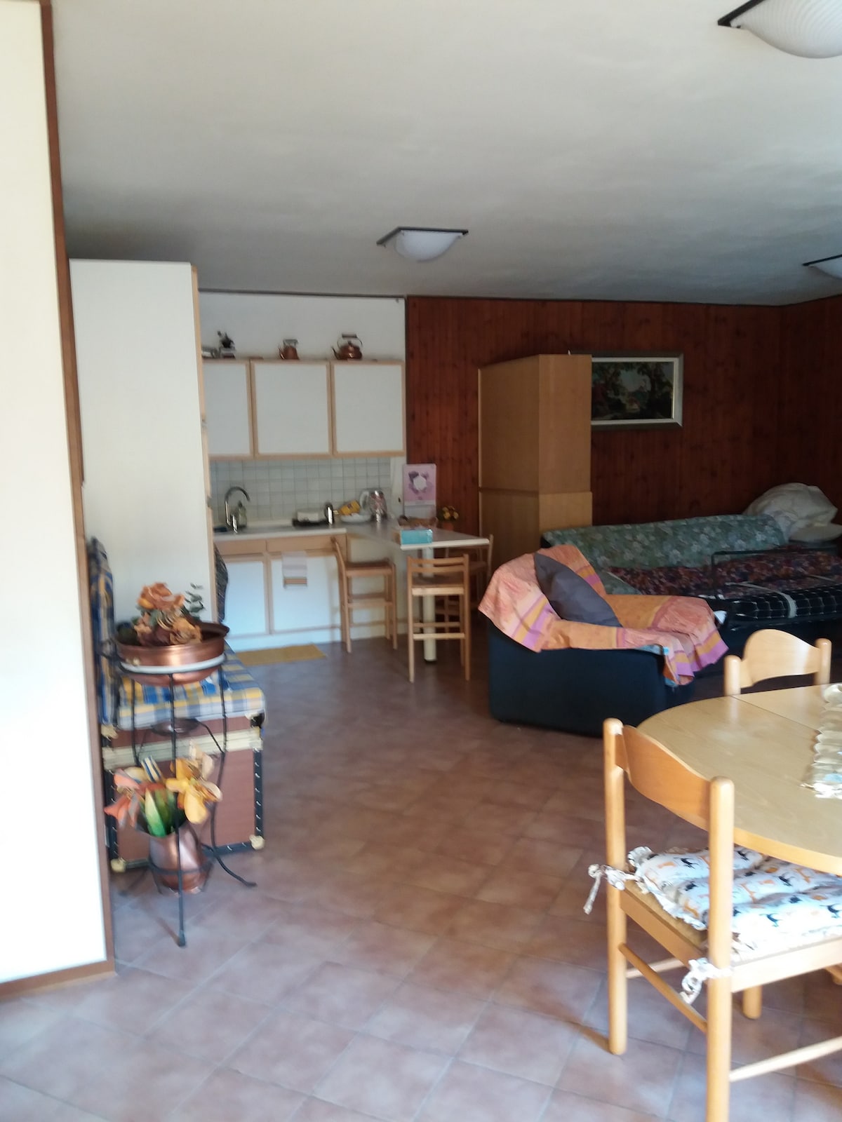 A living area is featured with a mix of seating options, including a black sofa and wooden chairs around a round table. The kitchenette is visible, displaying basic appliances and cabinetry. Light-colored walls contrast against wooden paneling, creating a warm and casual atmosphere.