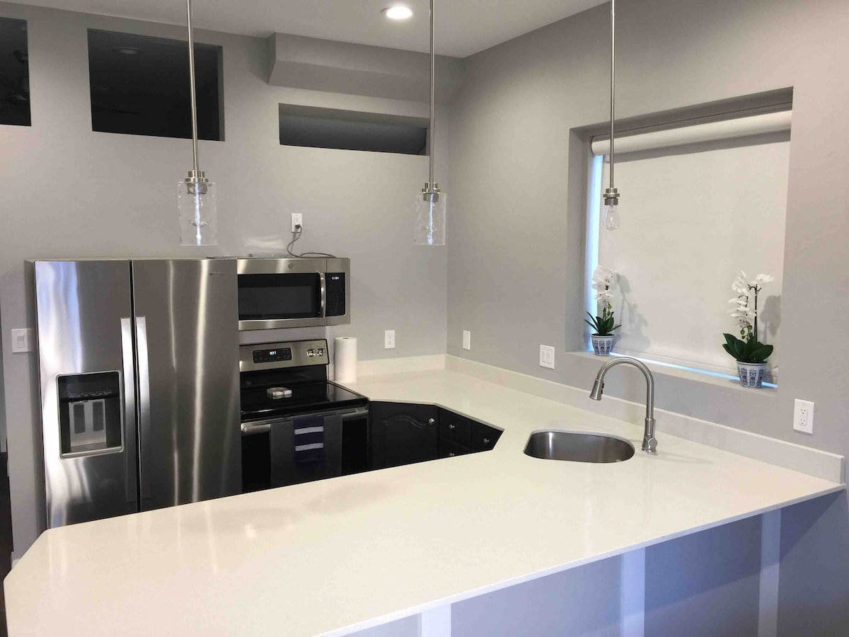 A modern kitchen area features stainless steel appliances, including a refrigerator and microwave. White countertops provide a clean workspace, complemented by contemporary pendant lighting. A window with a roller shade allows natural light to enter, and potted plants add a touch of greenery to the space.