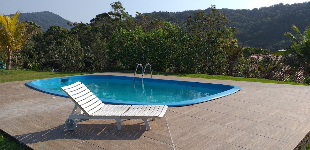 A tranquil outdoor pool is visible, framed by lush greenery and distant mountains. A single lounge chair rests beside the pool on a tiled patio, inviting relaxation amidst the serene natural surroundings.