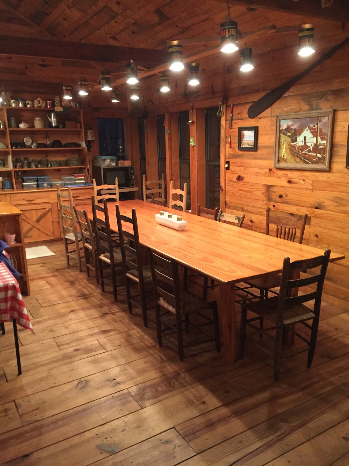 A spacious dining area features a long wooden table with seating for sixteen. Wooden chairs surround the table, enhancing the rustic charm. Soft lighting from hanging fixtures illuminates the room, highlighting the wooden walls and shelves filled with various kitchenware. A painting adds a decorative touch.