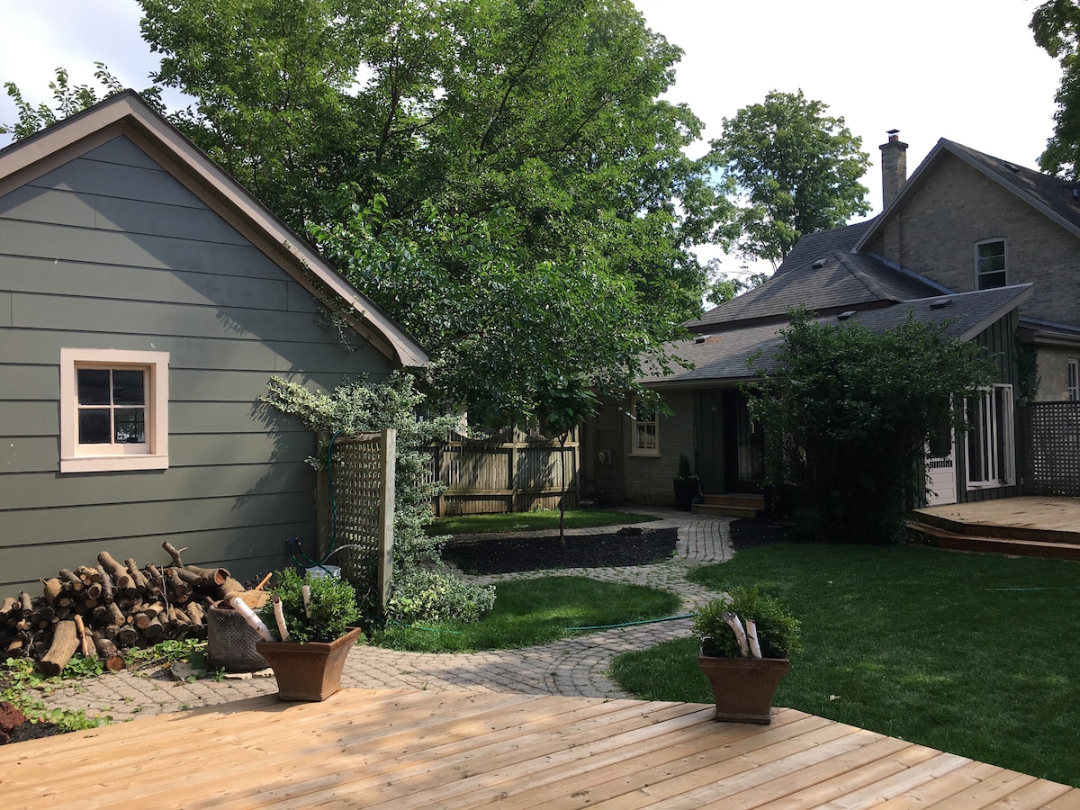 A landscaped outdoor area is visible, featuring a winding stone pathway that connects a green lawn to various gathering spaces. Two structures are seen, one with a gray exterior and the other with a light-colored façade. Floral plant pots are positioned on a wooden deck, contributing to the serene atmosphere.