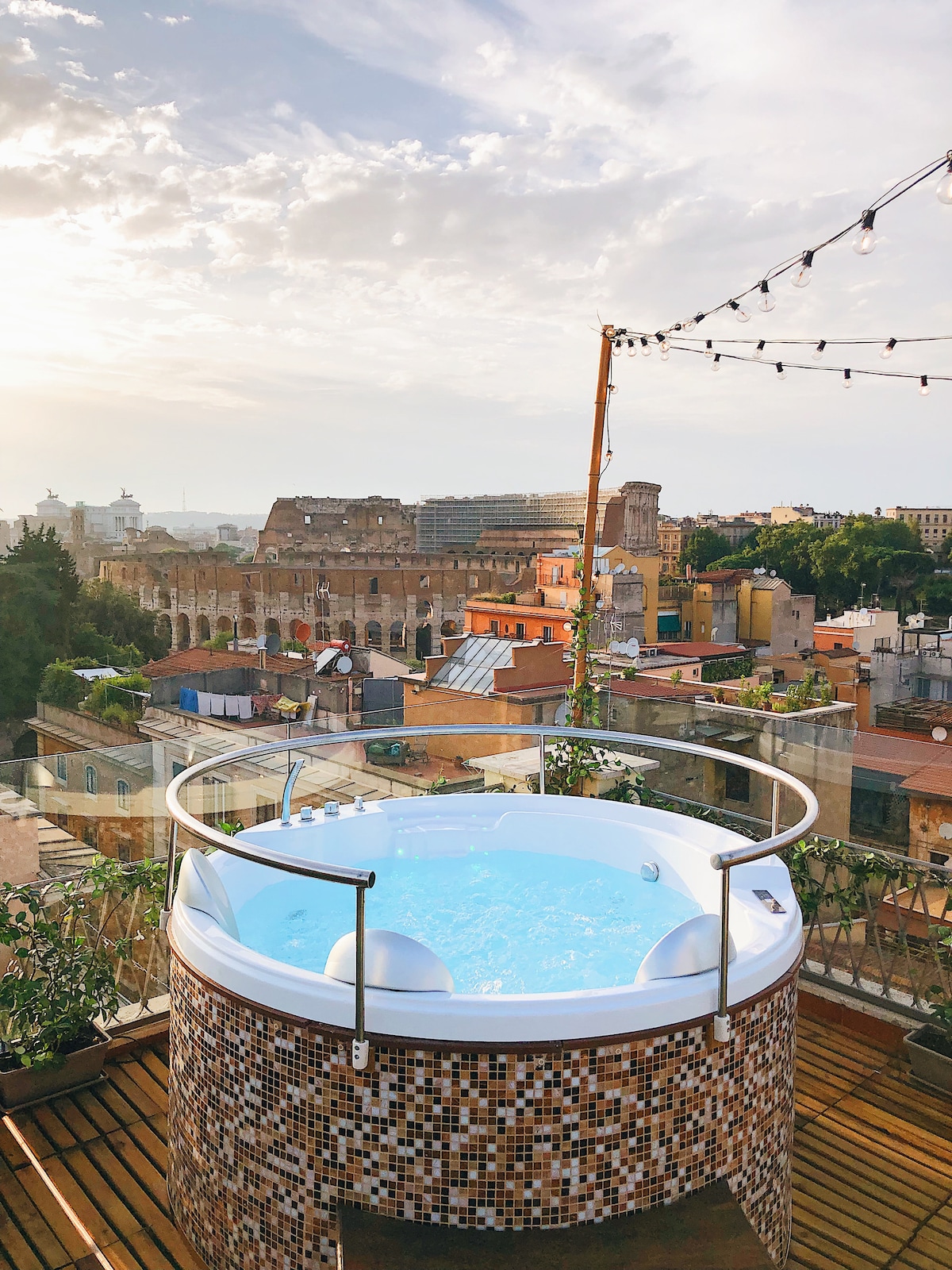 A private hot tub is situated on a wooden deck, surrounded by greenery. The view overlooks the rooftops of Rome, with the Colosseum visible in the distance under a sky filled with soft clouds and afternoon light.