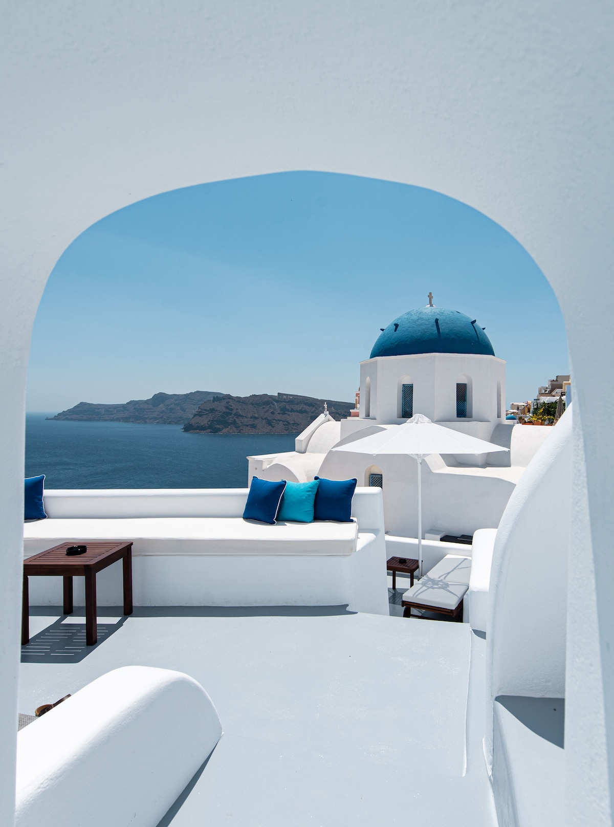 An arched entrance frames a stunning view of the caldera and ocean. A terrace is visible, featuring a small table and dark blue cushions arranged on white seating. An iconic blue dome is situated nearby, with an umbrella offering shade.
