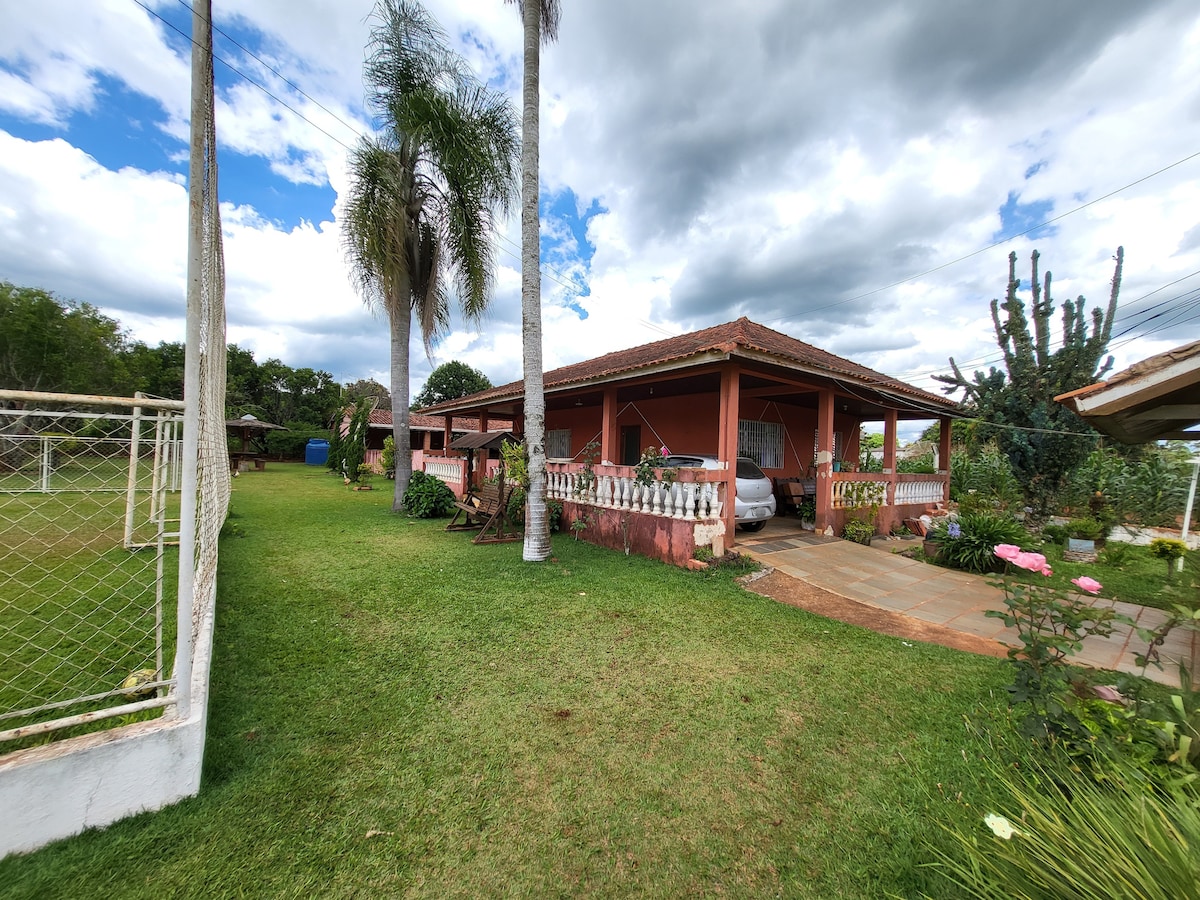 A spacious outdoor area features well-maintained grass and a mix of mature trees and shrubs. The house, designed with a covered porch, is framed by a clean walkway and nearby ornamental plants. Clouds drift across the sky, creating a serene backdrop.