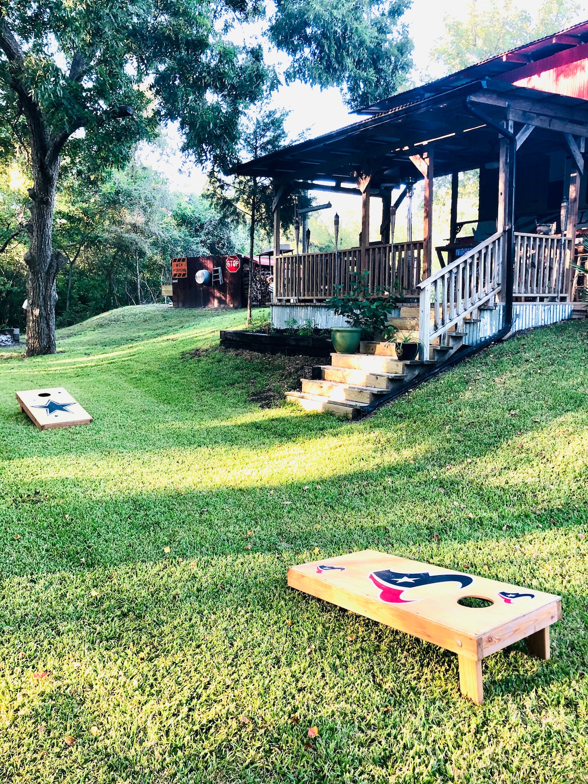 A well-maintained grassy area features a pair of cornhole boards, with a rustic structure visible in the background. Stairs lead up to a deck surrounded by trees, creating a welcoming outdoor space for games and relaxation.