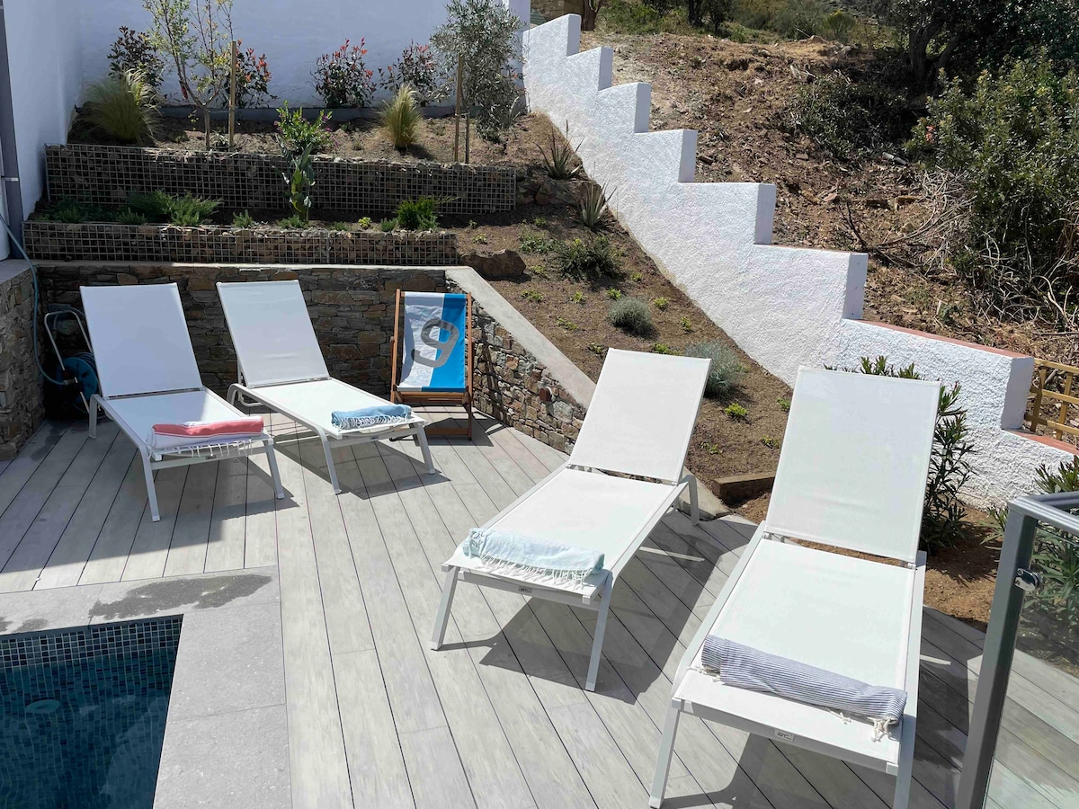 An outdoor space is presented featuring four lounge chairs arranged along a wooden deck. A saltwater pool is visible in the foreground, with a scenic hillside backdrop adorned with shrubs and plants. A staircase leads upwards beside the lounge area.
