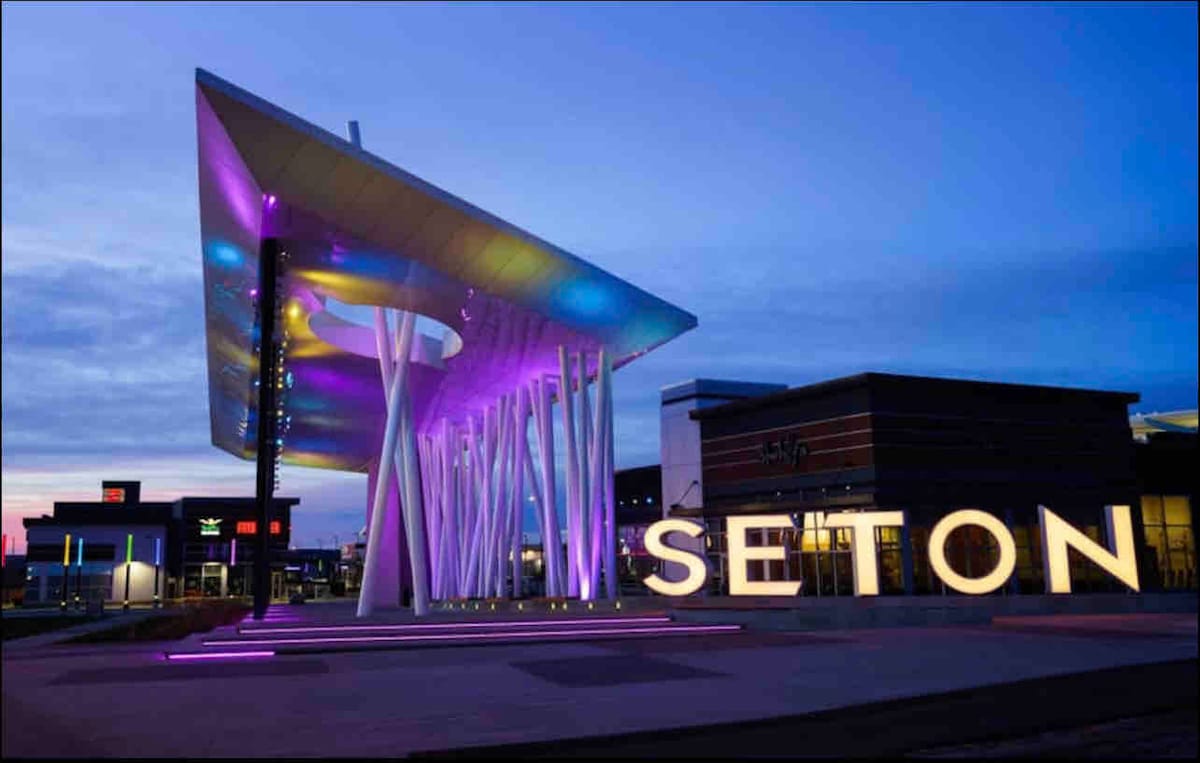 A modern structure with a large overhang and artistic white columns is illuminated by vibrant purple and blue lights. The word 'SETON' is prominently displayed in large letters, adding a distinctive touch to the entrance of the community, set against a dusk sky.