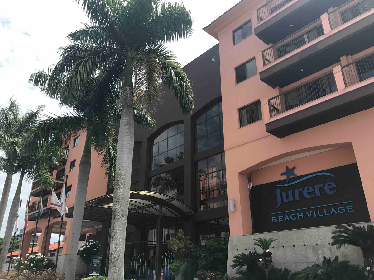 The exterior of Jurerê Beach Village is displayed, featuring a modern structure with large windows and multiple balconies. Palm trees frame the entrance, which is marked by a prominent sign. The building is painted in warm colors, complementing the inviting atmosphere.