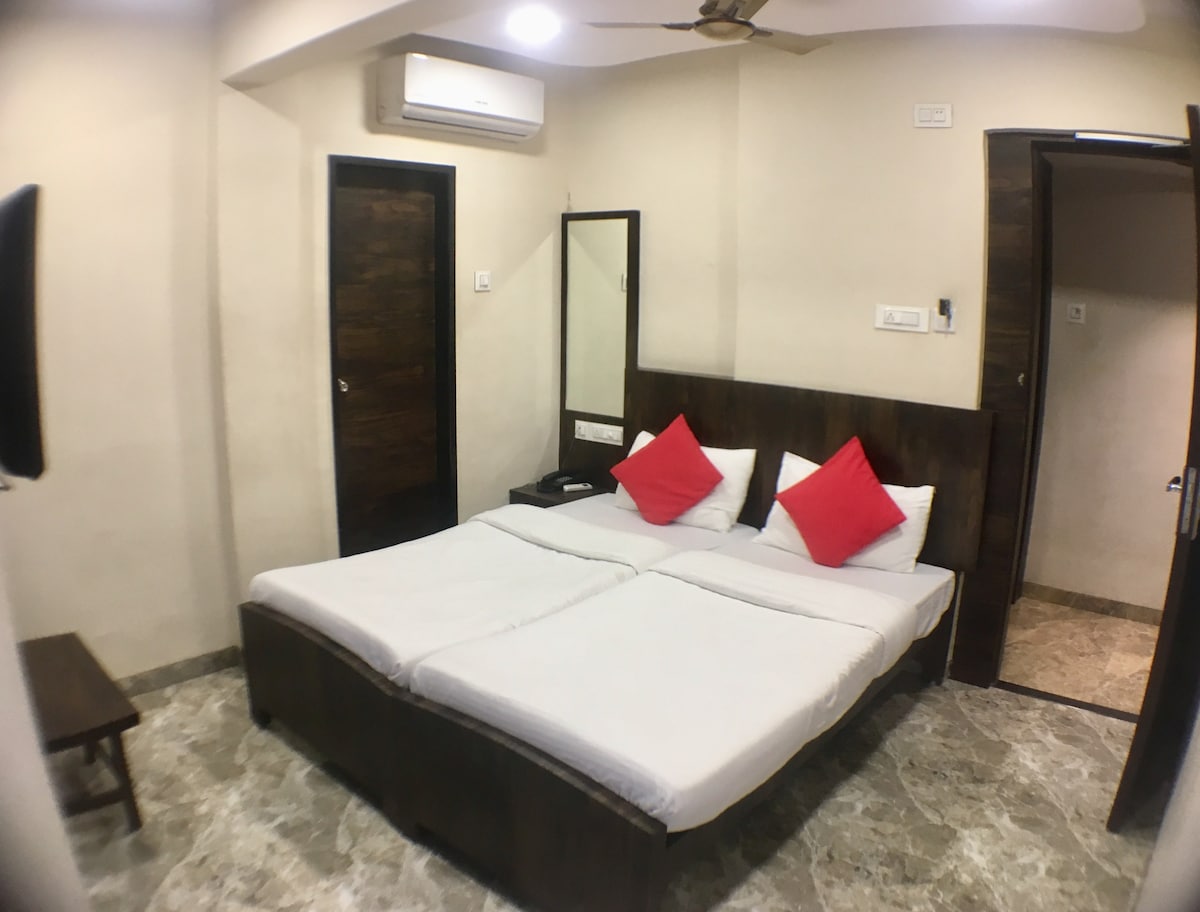 A well-furnished room features a spacious bed with light-colored bedding and contrasting red accent pillows. A mirror hangs above a bedside table, and air conditioning is installed near the ceiling. Natural light enters through a small window, and an open door leads to a bathroom.