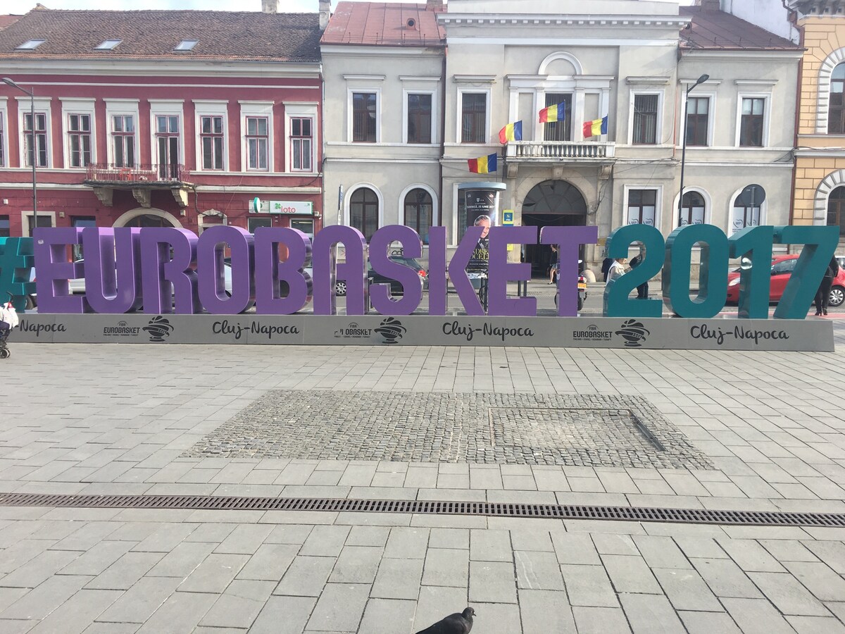 A colorful and large display features the hashtag '#EUROBASKET2017' in bold letters, set against a backdrop of historic buildings. The sign is situated on a paved square, creating a focal point for visitors and passersby.