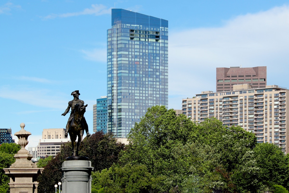 A historic statue of a mounted figure stands in a park, framed by lush greenery. In the background, modern skyscrapers rise against a clear blue sky, highlighting the contrast between the historic and contemporary architecture.