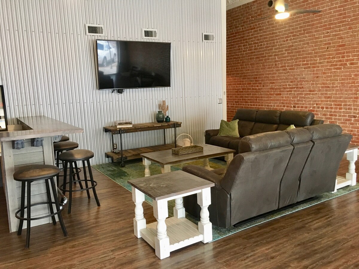 The spacious living area is furnished with a large brown sectional sofa, facing a 70” TV mounted on a textured white wall. A wooden coffee table is placed at the center, alongside two side tables. Bar stools are positioned at an adjacent counter, all set against exposed brick accents.