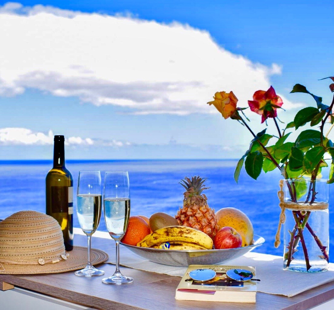 A table is set with a variety of fresh fruits, including pineapple, bananas, and oranges, alongside two glasses of sparkling wine and a bottle. A vase holds colorful roses, and a straw hat and sunglasses are placed nearby, all against a backdrop of the ocean and blue sky.