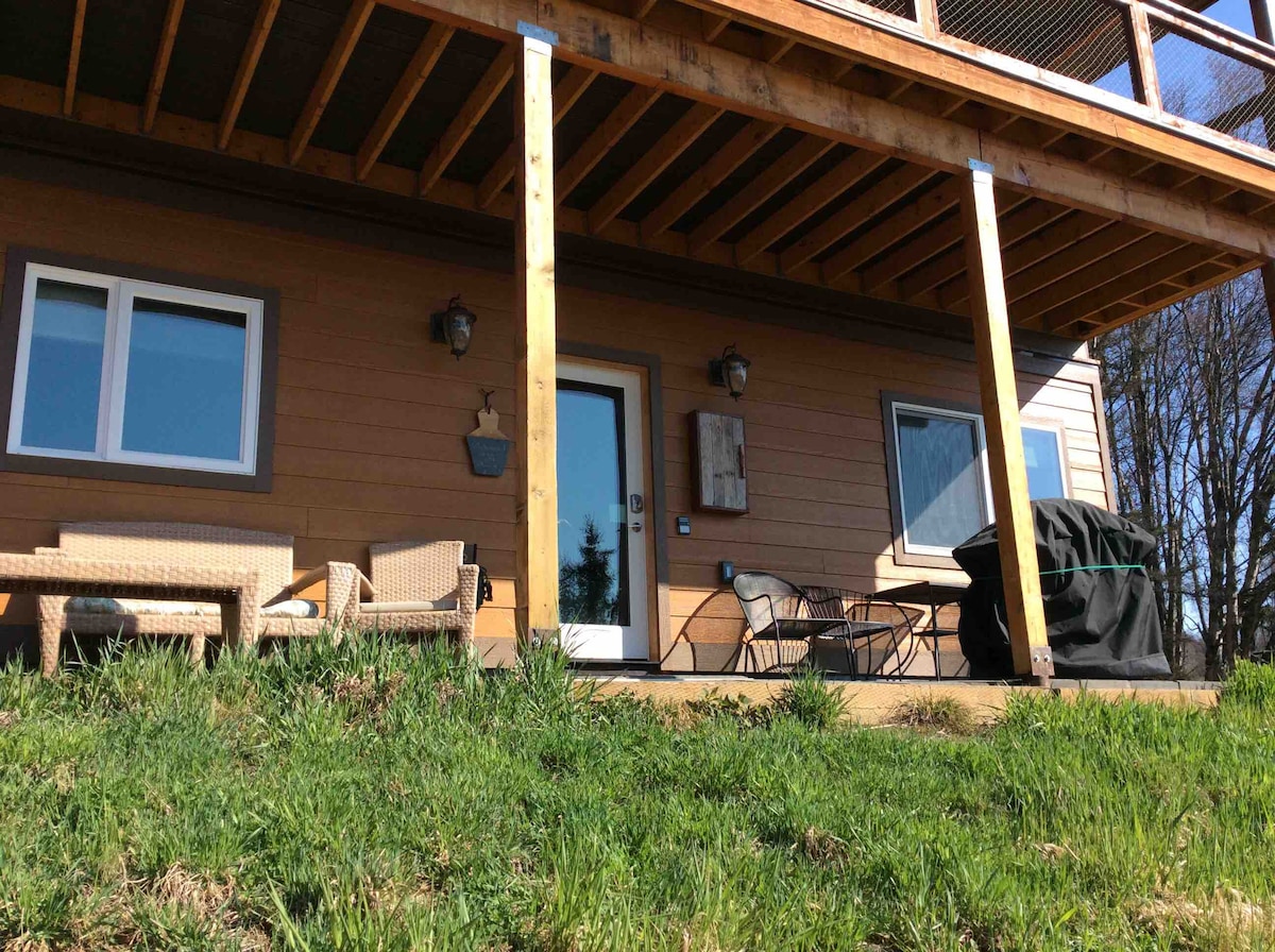 An exterior view showcases the ground floor of a newly built home, featuring a covered patio with outdoor seating arrangements. A grill is positioned nearby, and large windows allow for natural light to enter. The entrance is framed by wooden beams and a welcoming doorway.