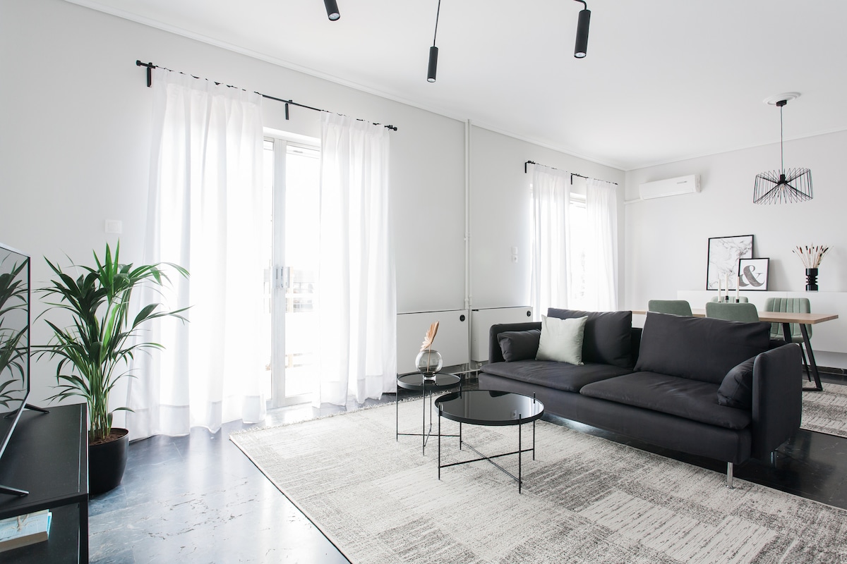 A modern living room is featured, showcasing a black sofa positioned across from a glass coffee table. Large windows with sheer white curtains allow natural light to enter. A minimalistic dining area is visible in the background, complemented by a green plant on the floor.