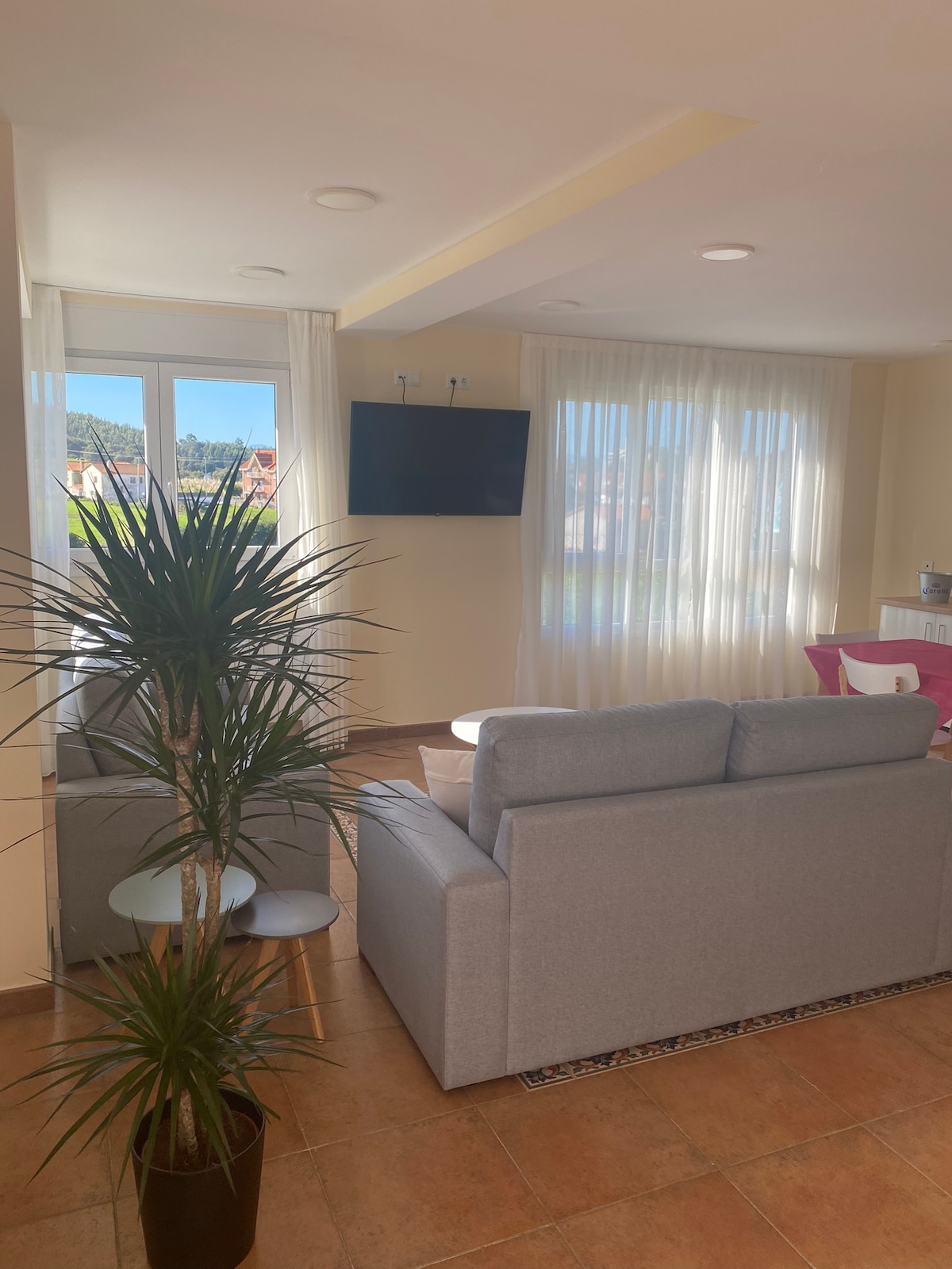 A modern living area features a light gray sofa positioned near a flat-screen television mounted on the wall. Large windows allow natural light to illuminate the space, while a potted plant adds a touch of greenery. Soft curtains frame the windows, creating an airy feel.