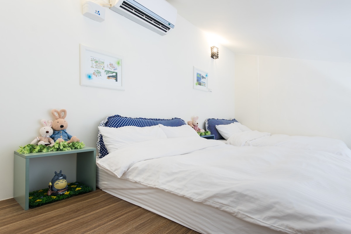 The sleeping area features two spacious beds topped with soft, white bedding. A pair of plush toys sits on each bed, accompanied by green side tables with decorative elements. Soft lighting from wall fixtures complements the bright, neutral walls, creating a cozy feel.