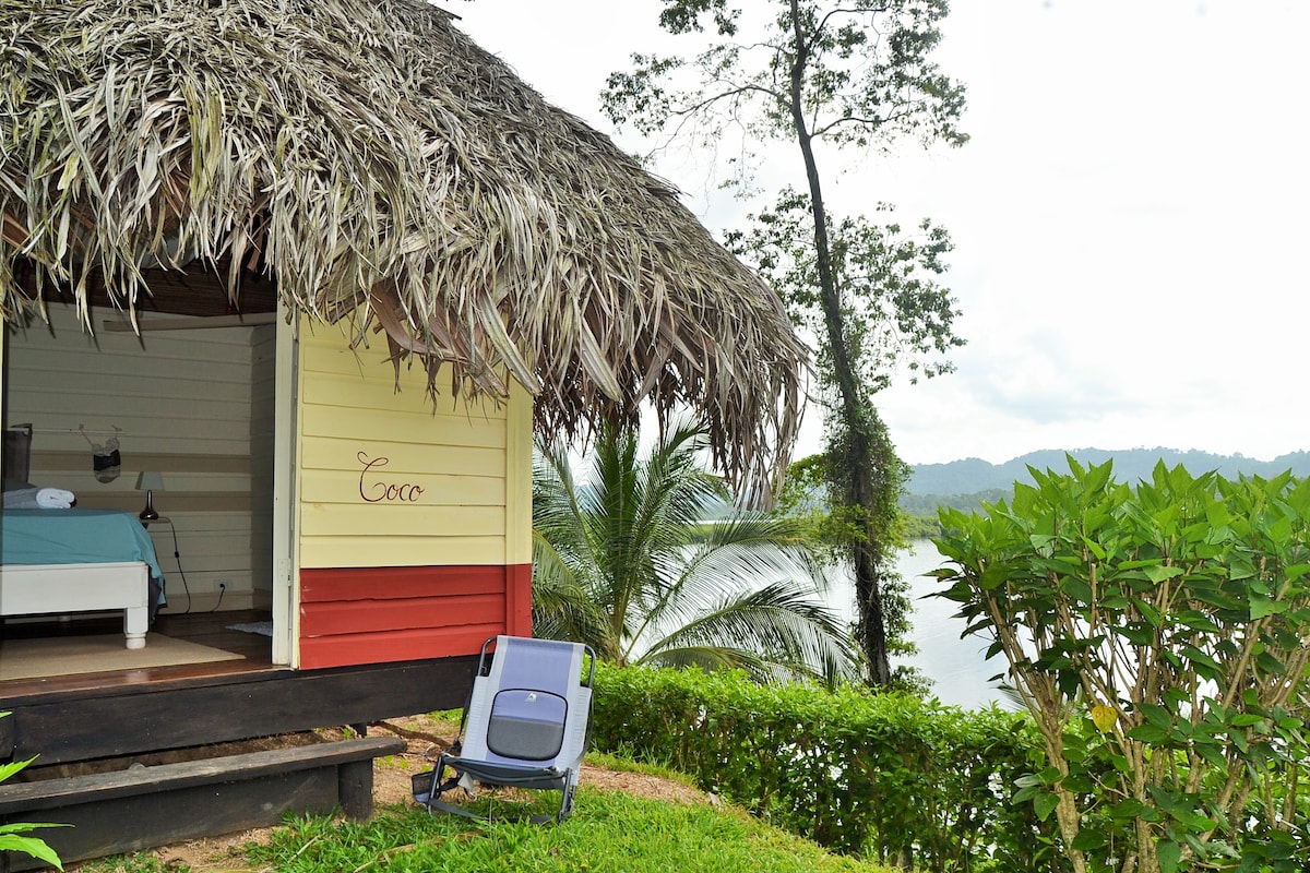 A bungalow with a thatched roof is positioned on elevated ground, overlooking a serene body of water. A single lounge chair is set on a natural surface, surrounded by vibrant greenery. The distant hills create a tranquil backdrop under a gently clouded sky.