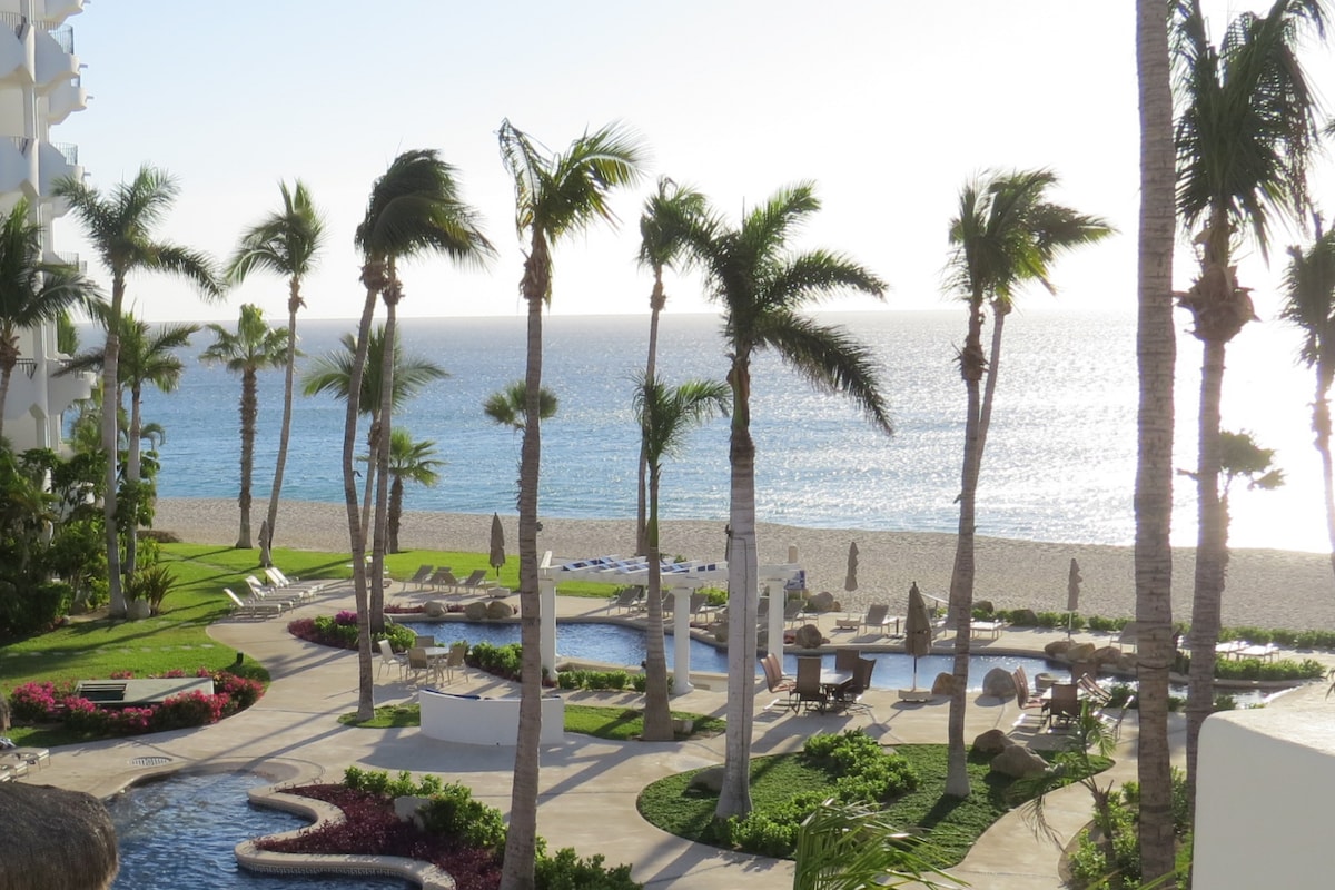 A serene outdoor area features palm trees swaying in the breeze, leading to a beach with gentle waves. Two swimming pools are visible, surrounded by lounge chairs and shaded by umbrellas. Vibrant flowers add color to the lush landscape, complementing the ocean's glimmering surface.