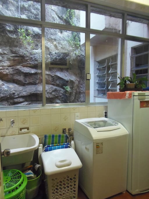 Área de serviço com tanque, máquina de lavar, geladeira, tábua de passar, ferro elétrico e varal.