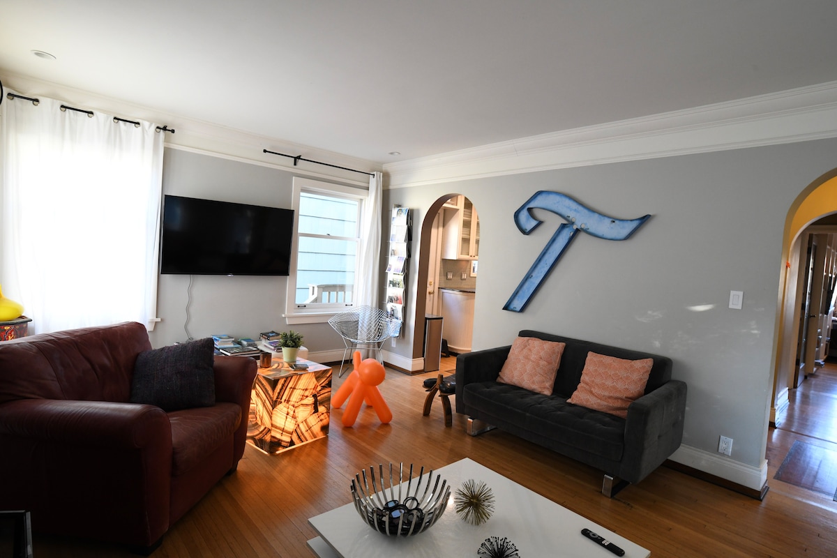 A spacious living area features comfortable seating with a leather couch and accent chairs. A large wall-mounted TV is visible, and natural light enters through the windows covered with sheer curtains. An artistic wooden table adds a touch of style to the hardwood floor.